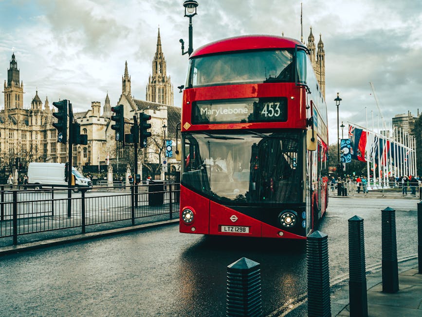 A red double-decker bus with route information displaying 'Marylebone' and the number '453' is parked on a wet street pavement in Marylebone, London. Behind the bus, there are historic Gothic-style buildings and the Houses of Parliament with its spires visible under a cloudy sky. To the right, a row of Union Jack flags hangs along a fence, and a few pedestrians and vehicles are present in the background. The bus is part of the public transport system in London, and the scene captures the urban environment typical of a busy home relocation or furniture transport area, with the bus likely used for logistics or moving services in the Marylebone area, as managed by Man With a Van Marylebone.