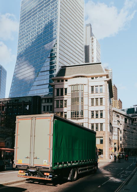 A large commercial moving truck with a green cargo container is parked on a city street in Marylebone, London, surrounded by tall modern glass skyscrapers and historic stone buildings. The truck is positioned near a sidewalk, with its rear doors closed, indicating the loading or unloading process typical in home relocation or furniture transport. The street appears to be busy, with a few pedestrians visible and some traffic in the background, under a clear sky with sunlight reflecting off the glass surfaces. The scene captures an urban environment where professional removals by companies such as Man With a Van Marylebone often facilitate efficient packing, moving, and transport of household furniture and boxes during house removals or relocation services.
