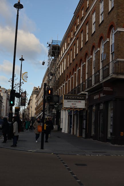 Street view in Marylebone showing a building with a brown brick façade and multiple floors, some with small balconies and white-trimmed windows. The building has scaffolding on the left side, indicating ongoing work. Pedestrians are walking along the pavement, some carrying shopping bags or backpacks, with a few waiting at a pedestrian crossing. A black van is partially visible on the right side, parked close to the curb where a crew from Man With a Van Marylebone is preparing for home relocation or furniture transport. Street lamps and decorative signs are also visible, adding to the typical urban environment. The scene captures the process of house removals, including loading or unloading activities, in a busy Marylebone street environment, with natural daylight illuminating the area.