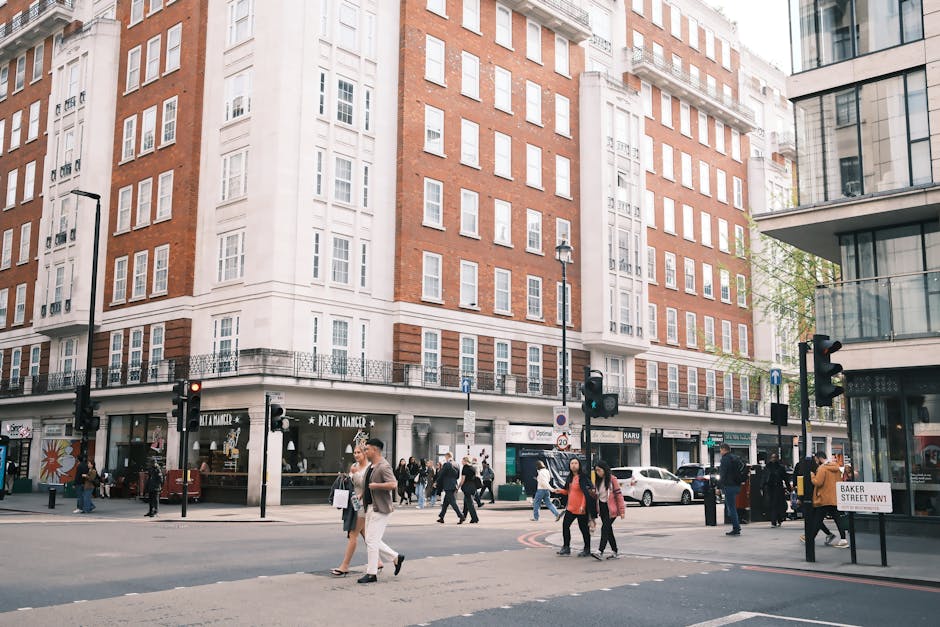A busy city street scene featuring the ground floor of a large, multi-storey building with a mix of red brick and white facade, housing various retail shops and cafes at street level. Pedestrians are crossing the road in front of the building, some carrying shopping bags or backpacks. Several cars are parked along the curb, and others are moving through the intersection. Utility poles and streetlights are visible, with traffic lights showing red for vehicles. The street sign indicates the location as Baker Street NW1, in Marylebone. The environment appears well-lit with natural daylight, and the scene captures typical urban movement associated with house removals or furniture transport, as part of a home relocation process facilitated by Man With a Van Marylebone, ensuring safe loading, transport, and unloading of household items.