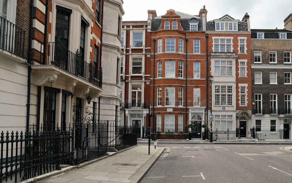 A row of Georgian-style terraced houses on a quiet street in Connaught Village, W1U, with ornate iron railings, white and red brick facades, and large sash windows. The pavement in front of the buildings is wide and clean, with a few trees and street lamps visible. The image shows a typical setting for house removals and home relocation services offered by Man With a Van Marylebone, with no moving activities or vehicles in view, but the surroundings reflect the urban environment suitable for furniture transport and packing and moving logistics in central London.