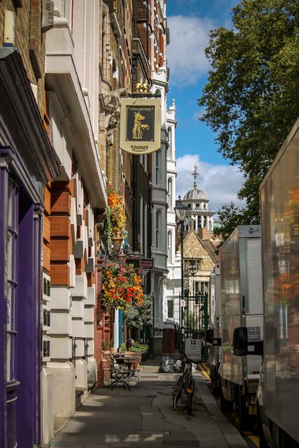A narrow street scene in Connaught Village, W1U, featuring a line of parked moving vans on the right side, with some of their side doors open for loading. Sidewalks are lined with multicoloured residential buildings, including a purple door and decorated window boxes with flowers. An outdoor seating area with small tables and chairs is visible along the pavement. Above, a street sign for 'Youngs' hangs from a building, and in the background, historic church domes and spires can be seen beneath a partly cloudy sky. The scene captures the typical setting for home relocation services, where furniture, boxes, and packaging materials are loaded onto transport vehicles, with some items such as cardboard boxes, wrapped furniture, and plastic packaging visible near the vans, illustrating the process of packing and moving in a busy urban environment. Man With a Van Marylebone provides comprehensive removals within this area, supporting furniture transport and home relocation logistics.