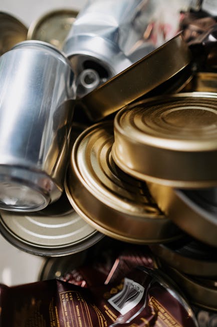 A close-up view of a pile of empty aluminum cans, some with a silver finish and others with a gold-colored coating, stacked haphazardly on top of each other on a surface. In the background, partially visible, are additional cans and packaging materials. This arrangement suggests the process of collecting recyclable materials, which may be part of the packing and disposal tasks involved in house removals or a home relocation. The lighting highlights the metallic surfaces, emphasizing their reflective quality. Man With a Van Marylebone, a specialist in removals and moving services, often handles the sorting and transport of recyclables during the moving process, ensuring proper waste and recycling rules are followed post-move as explained on their website.