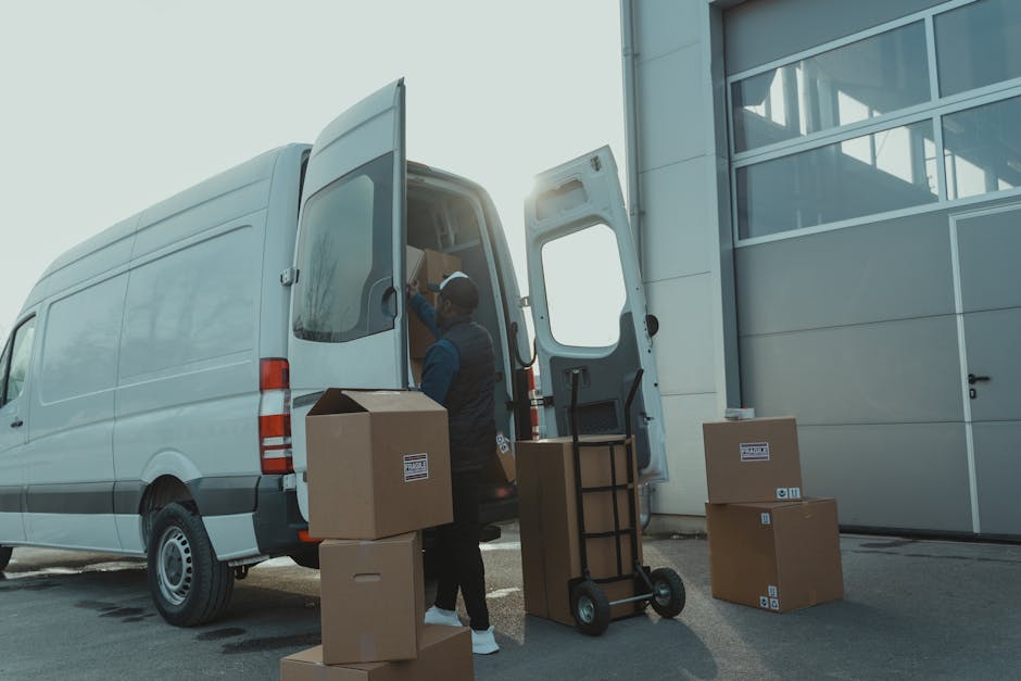 A professional mover from Man With a Van Marylebone stands beside a white moving van parked outside a building with a large glass door. The mover, dressed in dark clothing and a cap, is loading or arranging cardboard boxes into the rear cargo area of the van, which is open and reveals packing materials and other boxes inside. Several additional cardboard boxes, sealed and labelled, are positioned on the pavement near the van, with some stacked and others resting on the ground. A black trolley with a handle is placed next to the boxes for transporting items, and plastic wrapping and packing supplies are nearby. The scene captures the loading process during a home relocation, emphasizing careful handling and organized packing for furniture transport and moving services, consistent with safe and efficient logistics for house removals in the Marylebone area. Bright daylight illuminates the scene, highlighting the tactical positioning of boxes and equipment used in professional packing and moving tasks.