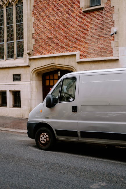 A grey cargo van parked on a street adjacent to a historic brick building with large, arched windows and a stone doorway. The van, used for home relocation or furniture transport, is positioned with its front facing right and features black side mirrors and wheels. Inside the cargo area, visible through an open side door, are several cardboard boxes, wrapped furniture pieces, and packing materials, indicating an ongoing loading or unloading process. The van is parked on a paved road, with the building’s exterior showing stone detailing and a security camera mounted near the top corner. This scene reflects the logistics involved in house removals, with careful loading of packing and moving supplies into the vehicle, supported by a professional service such as Man With a Van Marylebone, which handles parcel and furniture transport efficiently within Marylebone and the Portman Estate area.