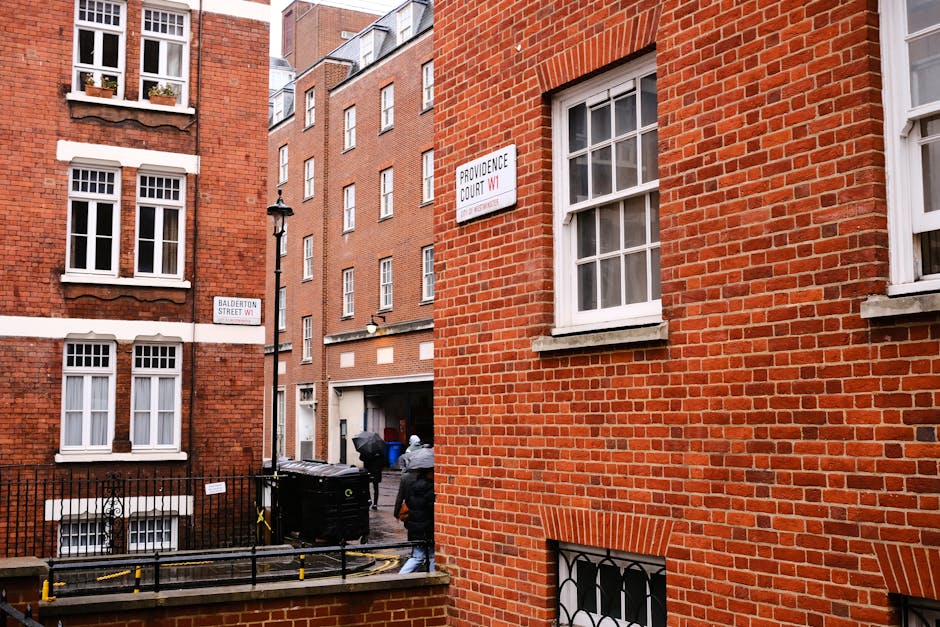 The image depicts a street scene in Marylebone, London, with a view of red brick residential buildings featuring white-framed sash windows. Two individuals are engaged in loading or unloading items into a black van parked on the pavement, with a few cardboard boxes and plastic-wrapped furniture pieces visible near the vehicle. One person is wearing a grey jacket and is walking towards the van while the other is partially visible behind a trash bin. The van is positioned in a loading area adjacent to the buildings, with a residential gate and some yellow barriers on the pavement nearby. Street signs indicating 'Balderton Street W1' and 'Providence Court W1' are attached to the buildings, contributing to the urban context. The scene is well-lit with natural daylight, emphasizing the typical architecture of a London home relocation, where furniture transport and packing may be underway, and a professional removals service like Man With a Van Marylebone is likely managing the logistics.