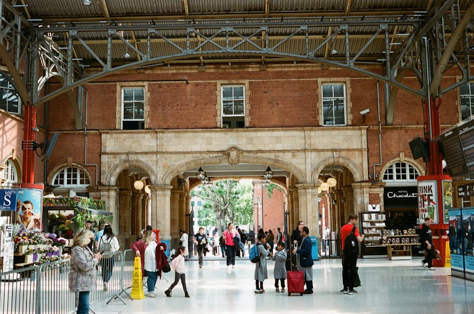Inside a busy train station with a high arched metal ceiling and large brick walls, several people are engaged in various activities related to a home relocation or travel. In the foreground, a group of individuals, including adults and children, are waiting or walking, some carrying luggage or backpacks. Near a small flower stand on the left, a woman is holding a smartphone, while another person with a child is standing nearby. To the right, a staff member in a black uniform appears to be assisting, standing next to a small shop selling chocolates and souvenirs, with shelves displaying items behind a black counter. Several cardboard boxes and plastic-wrapped packages are scattered around, indicating packing or transport preparations. The scene captures the logistical efforts involved in moving or travel arrangements, with the station environment highlighting the process of furniture transport and packing prior to a house removal or relocation service, as provided by Man With a Van Marylebone.