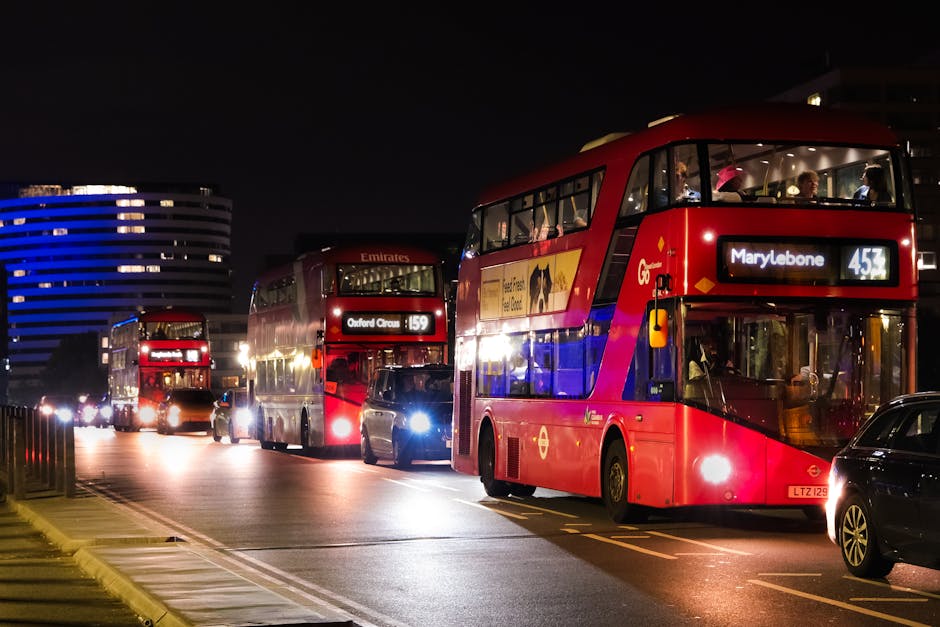Nighttime street scene featuring multiple double-decker buses illuminated by interior and exterior lights, parked along a street in Marylebone. The foremost bus displays route number 453 to Marylebone, with the destination sign clearly visible, while another bus further back shows route 159 to Oxford Circus. Several cars and a van are parked nearby, with some vehicle headlights on, reflecting off the wet road surface. The background includes modern, curved office buildings with blue lighting accents and lit windows, creating a vibrant urban atmosphere. The scene captures a typical busy evening during home relocation or furniture transport logistics, with buses and vehicles possibly used for moving or transporting household items, supported by companies like Man With a Van Marylebone. The lighting emphasizes the operational nature of the scene, highlighting transport and loading activities associated with moving services in the Marylebone area.