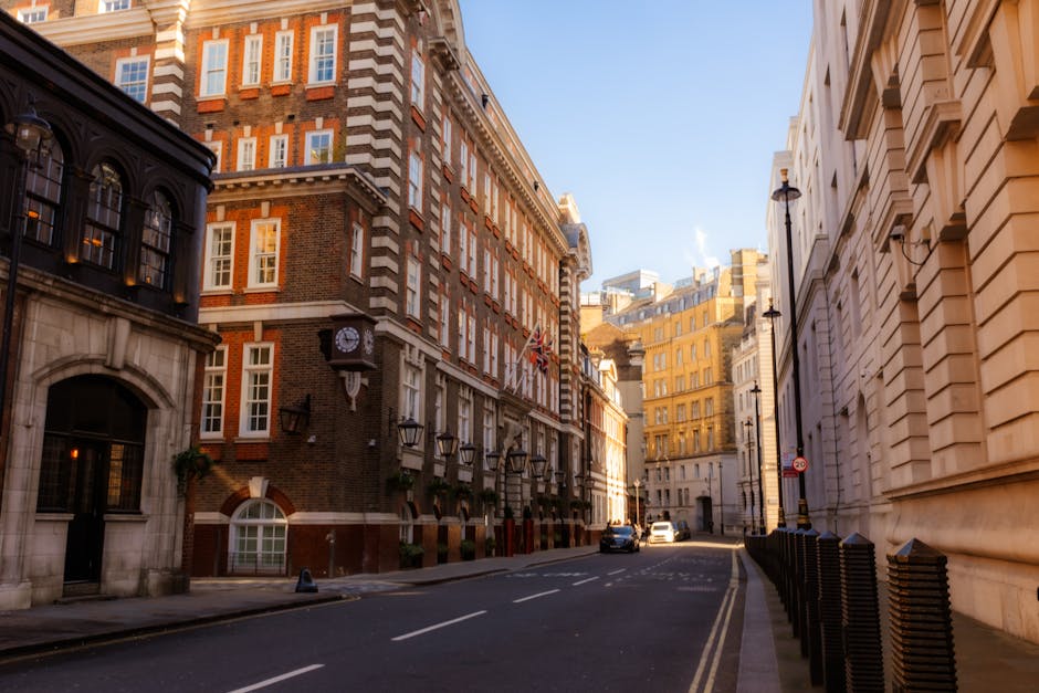 Photograph of a narrow street in Marylebone, Westminster, showing multi-storey historic buildings with brick and stone facades, some featuring decorative cornices and large windows. Several black lampposts line the pavement, and a few cars are parked along the kerb. The sky is clear with bright sunlight illuminating the scene. In the foreground, the street appears to be part of a home relocation or furniture transport process, with a van likely nearby for removals services. The environment suggests a quiet, urban area suitable for house removals, with the street providing space for loading or unloading furniture and boxes. The presence of a clock on one building indicates a traditional, well-maintained neighborhood often associated with professional removals such as those offered by Man With a Van Marylebone, operating within Westminster Council permits for residential moves.