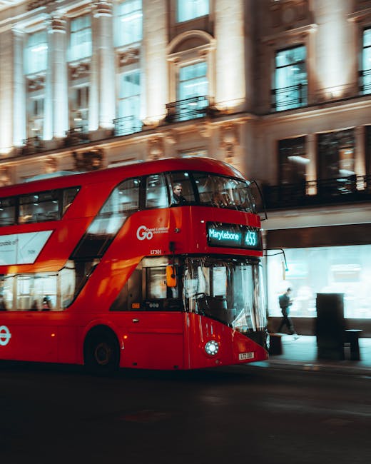 A red London double-decker bus, displaying route information for Marylebone on its electronic sign, is traveling along a city street during daytime. The bus is contemporary in design with large tinted windows, and it appears to be in motion, indicated by slight motion blur. In the background, historic multi-storey buildings with decorative facades and balconies are visible, illuminated by natural light. The street is populated with pedestrians, some walking on the sidewalk. To support house removals and furniture transport logistics, nearby is an area with open building doorway and pavement, where a person is seen carrying a cardboard box, and packing materials like plastic wrap and cardboard boxes might be hinted at nearby. This setting reflects an urban environment suitable for home relocation activities, with Man With a Van Marylebone providing removals services and supporting efficient furniture transport and packing during residential moves.